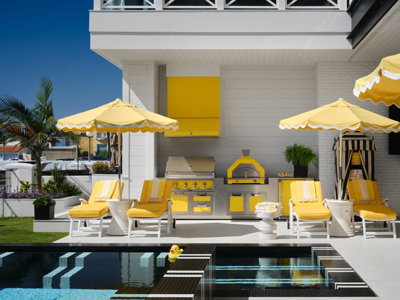 Sunny poolside patio with yellow and white loungers, umbrellas, and a modern outdoor kitchen. A vibrant, cheerful ambiance is accentuated by a rubber duck in the pool.
