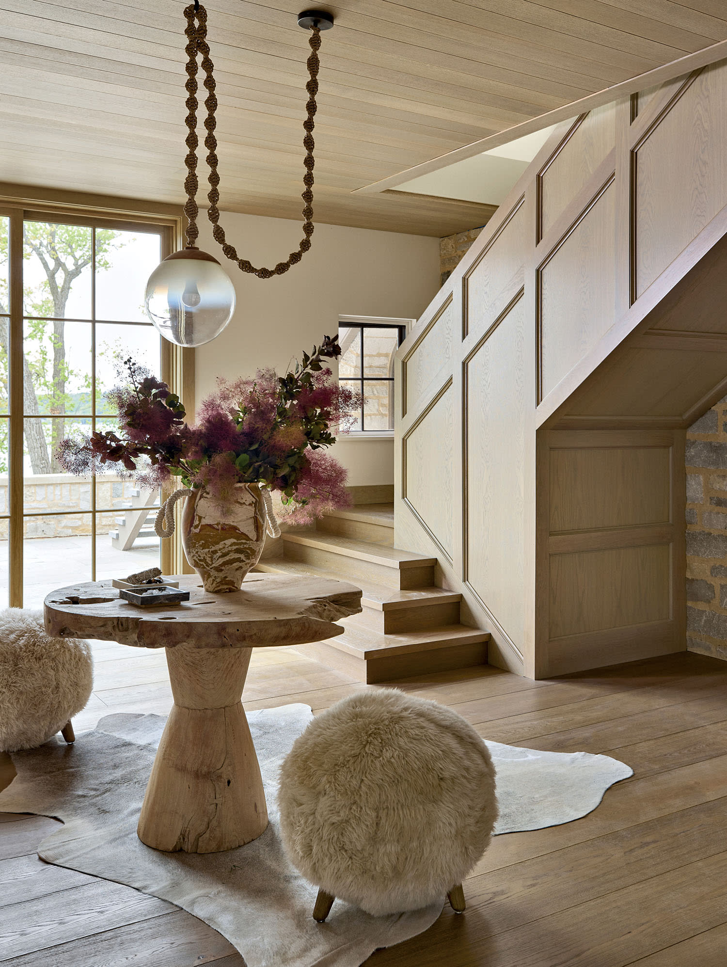 Cozy rustic interior with wooden stairs, a round wooden table, fluffy stools, and a vase of purple flowers. Natural light streams from large windows.