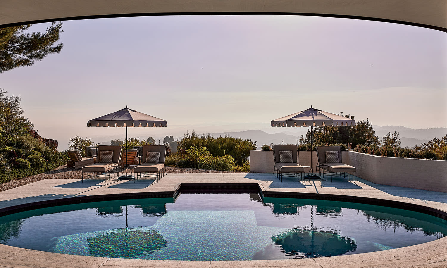 Luxury patio with a round pool reflecting two umbrellas. Lounge chairs and lush greenery overlook distant mountains under a serene sky. Peaceful ambiance.
