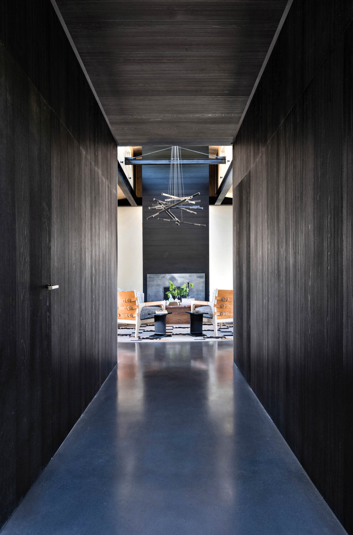 Sleek hallway with dark walls leading to a bright room featuring modern chairs, a plant on a table, and a sculptural ceiling light, creating a contemporary feel.