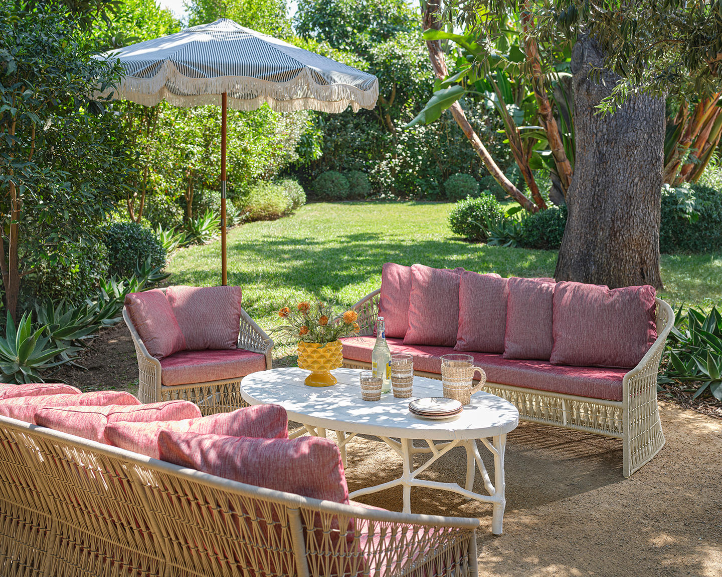 Outdoor patio with wicker furniture, pink cushions, and a white umbrella. Surrounded by lush greenery, the scene exudes a tranquil, relaxed vibe.