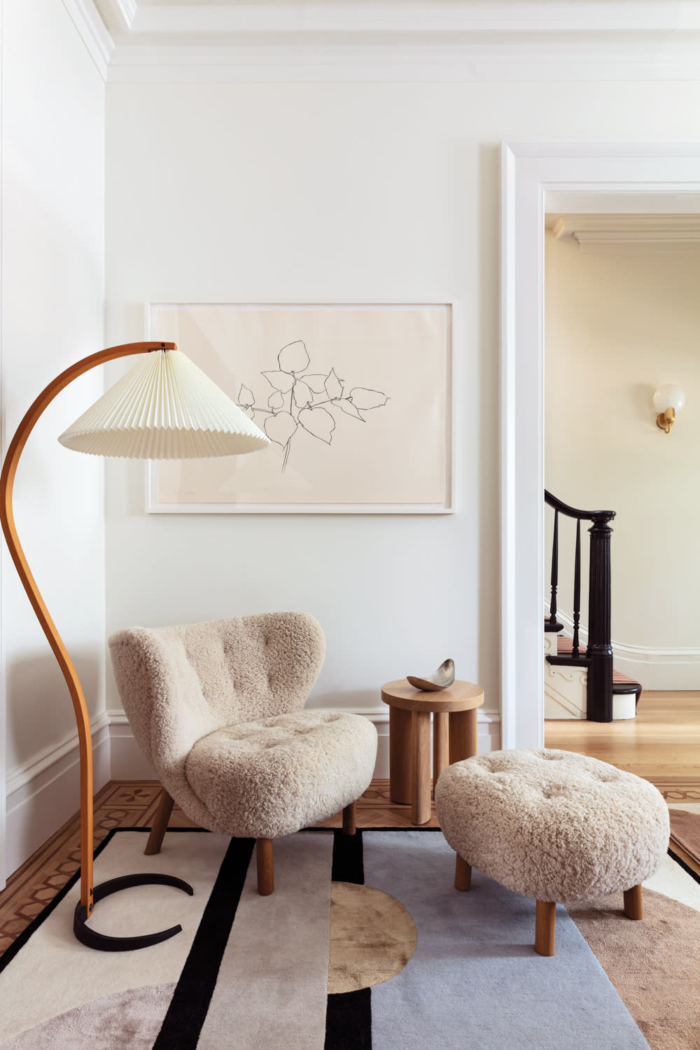 Cozy corner with a plush beige chair and ottoman, wooden lamp, abstract rug, and minimalist art. The space exudes warmth and elegance.