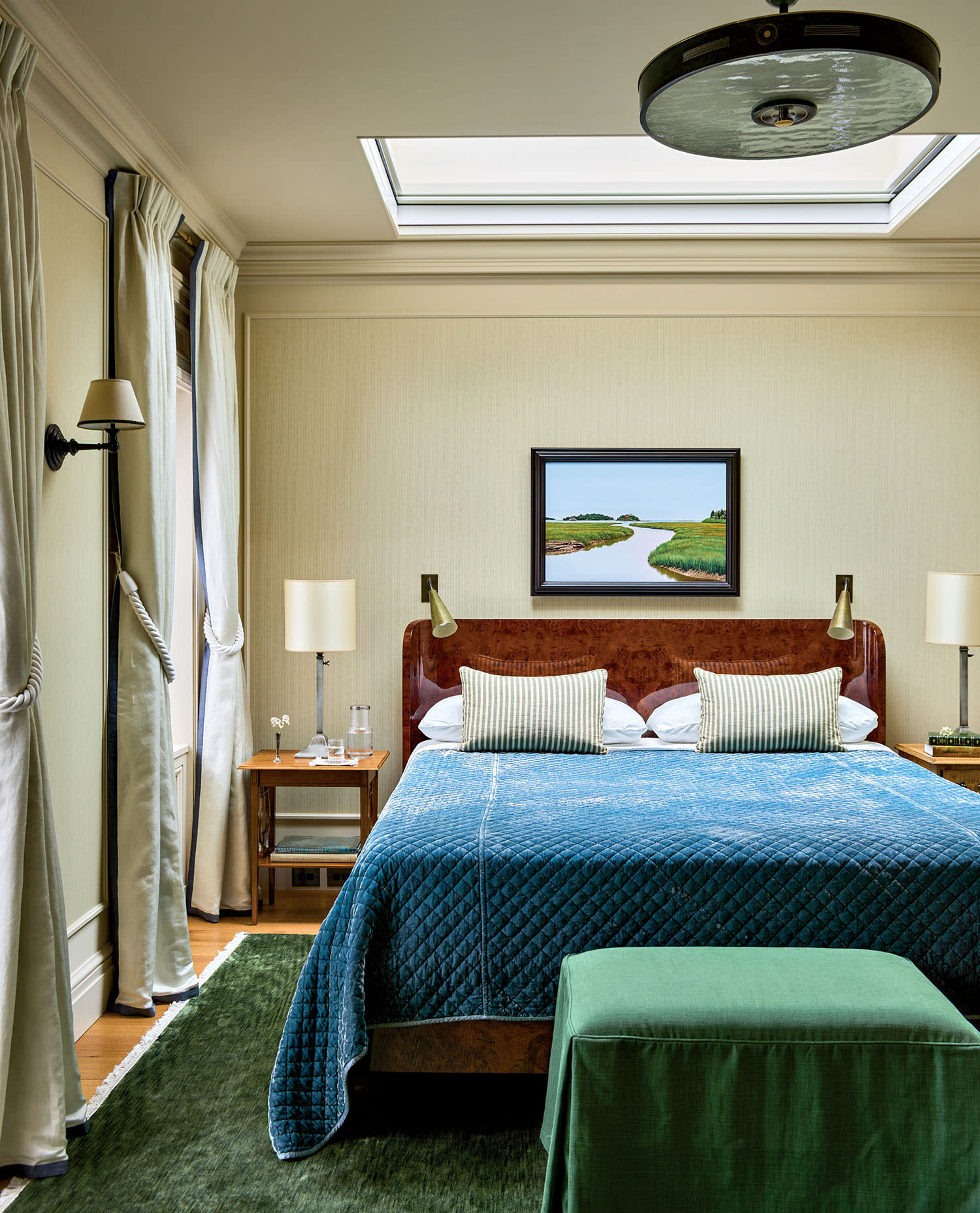 Elegant bedroom with a skylight, featuring a blue quilted bed, green ottoman, and landscape painting. Soft lighting and neutral curtains create a serene atmosphere.