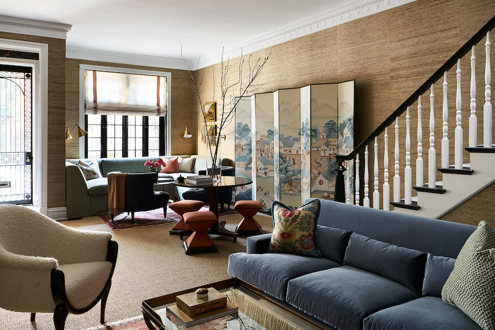 Large living room with natural light for front door and window. Neutral color grass cloth covers the walls and natural carpeting. Navy velvet sofa, orange stools surrounding table and decorative screen adds color and texture to room.