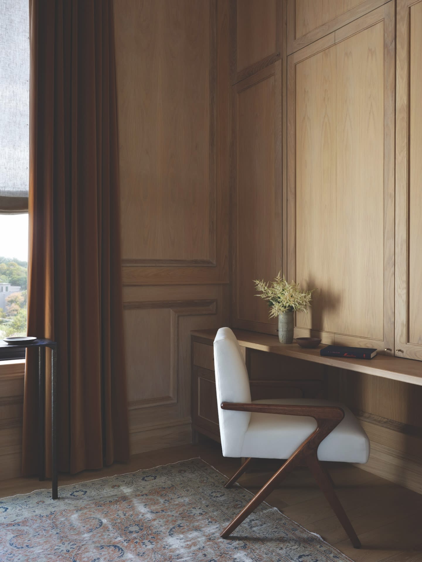 Cozy study with a wooden desk, white modern chair, and a beige vase with flowers. Soft light filters through brown curtains, creating a warm atmosphere.