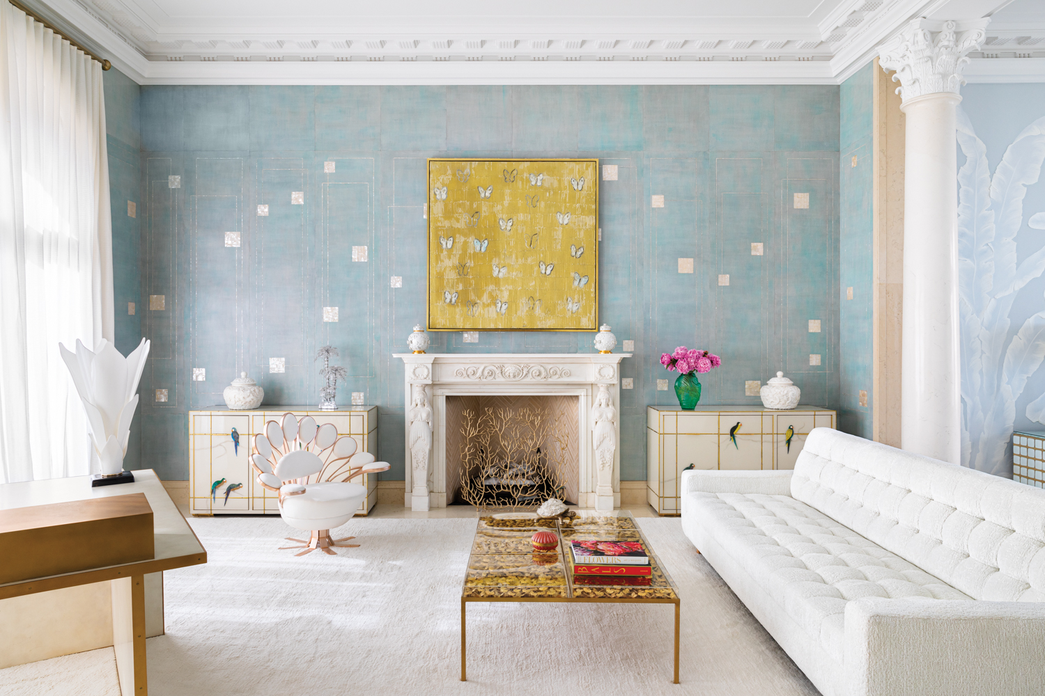 sitting area with light blue patterned wallcovering, gold artwork and white consoles, fireplace surround, sofa and rug