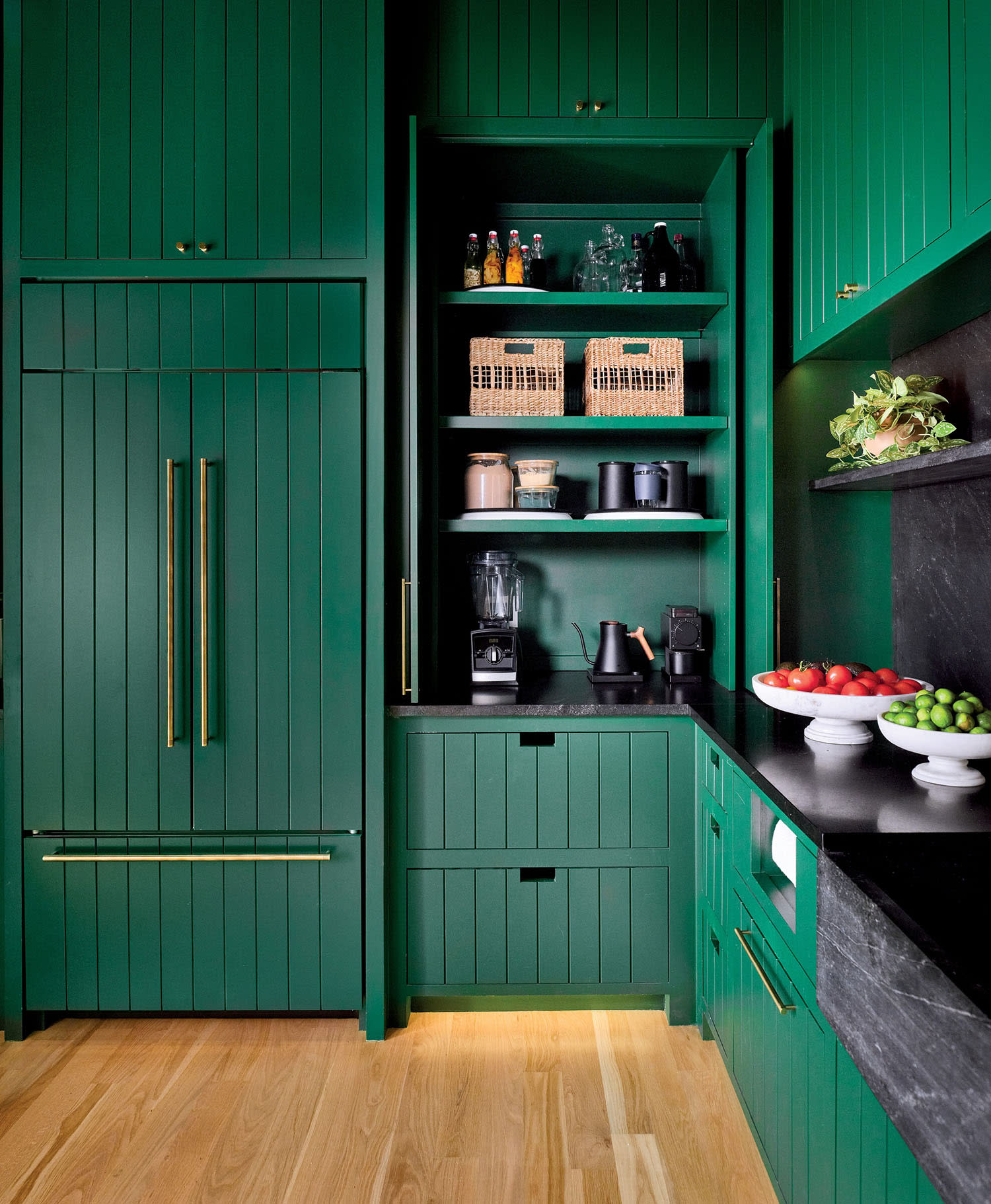 Green kitchen with floor-to-ceiling cabinets, brass handles, and a sleek black countertop. Open shelves hold baskets, bottles, and appliances.