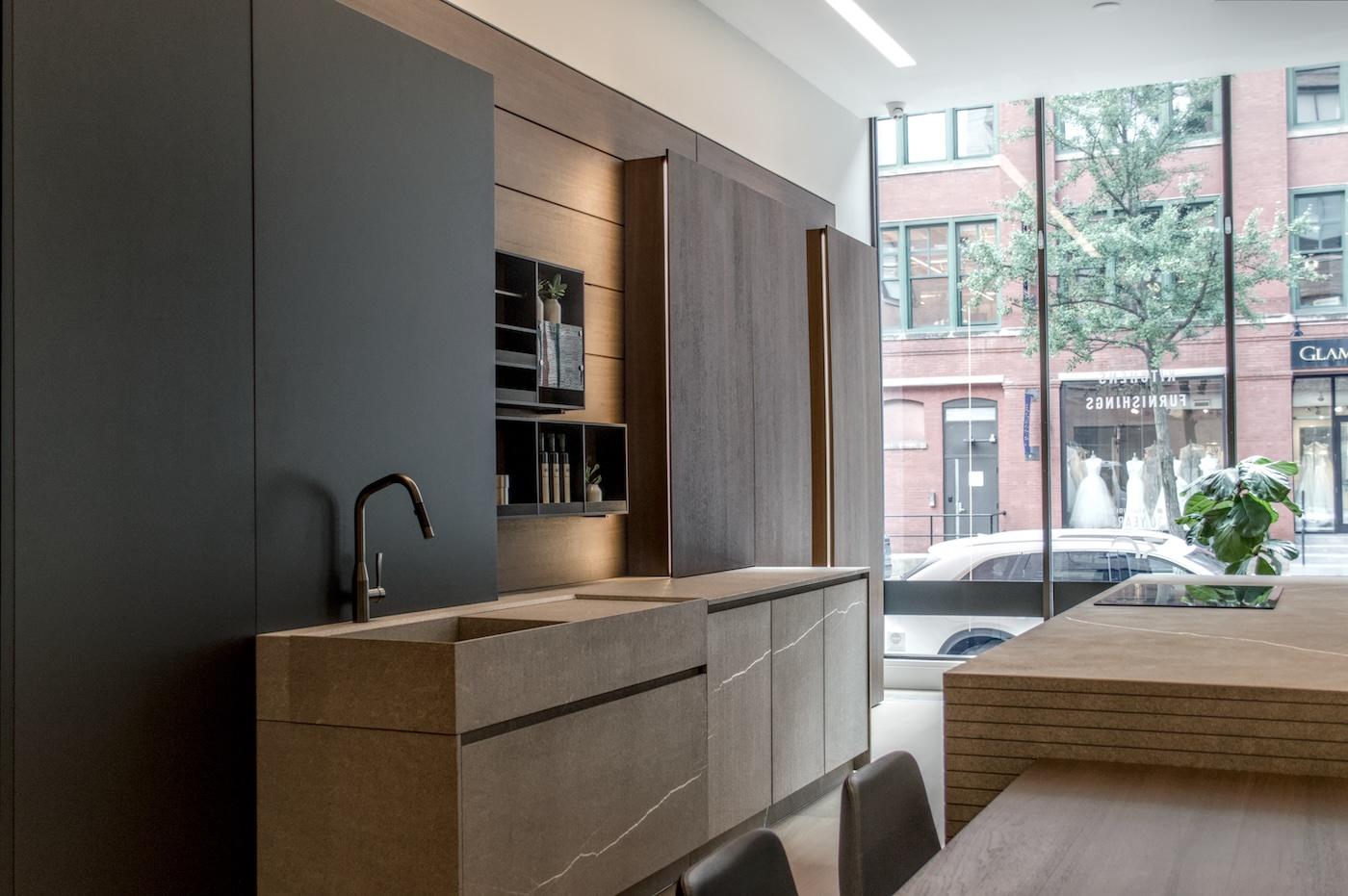 Modern kitchen with sleek dark cabinets, stone countertops, and a black faucet. Large windows reveal a red brick building and tree outside.