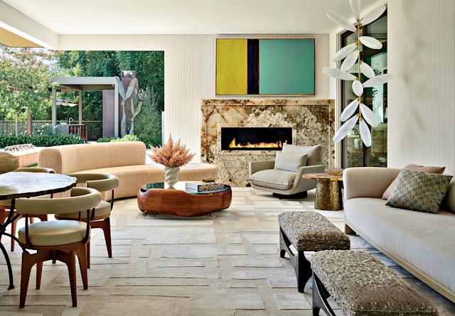 Stylish living room with modern furniture, a marble fireplace, abstract art, and large windows revealing a lush garden. The atmosphere is cozy and inviting.