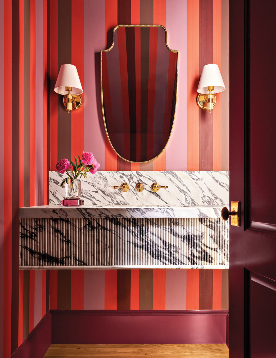 Modern bathroom with a marble sink against striped red and pink walls. A gold-framed mirror and wall sconces create an elegant, vibrant atmosphere.