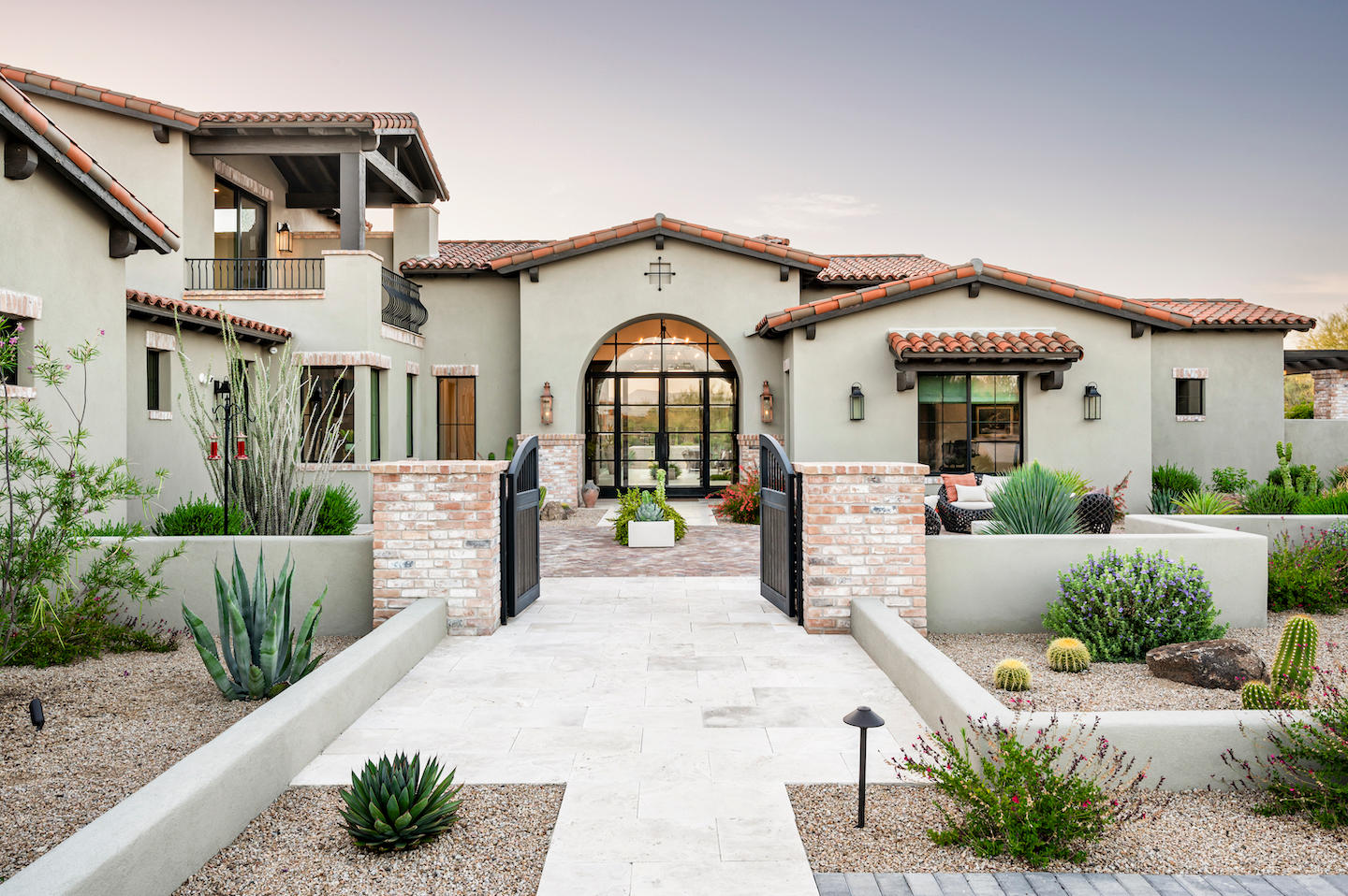 Spanish-inspired villa entrance featuring sage-green stucco walls, red barrel-tile roofs, and an arched glass front door framed by rustic brick pilasters and twin black wrought-iron gates. A light stone walkway leads through xeriscape beds of agave, barrel cacti, and desert shrubs, setting a warm, welcoming vibe beneath a pastel evening sky.
