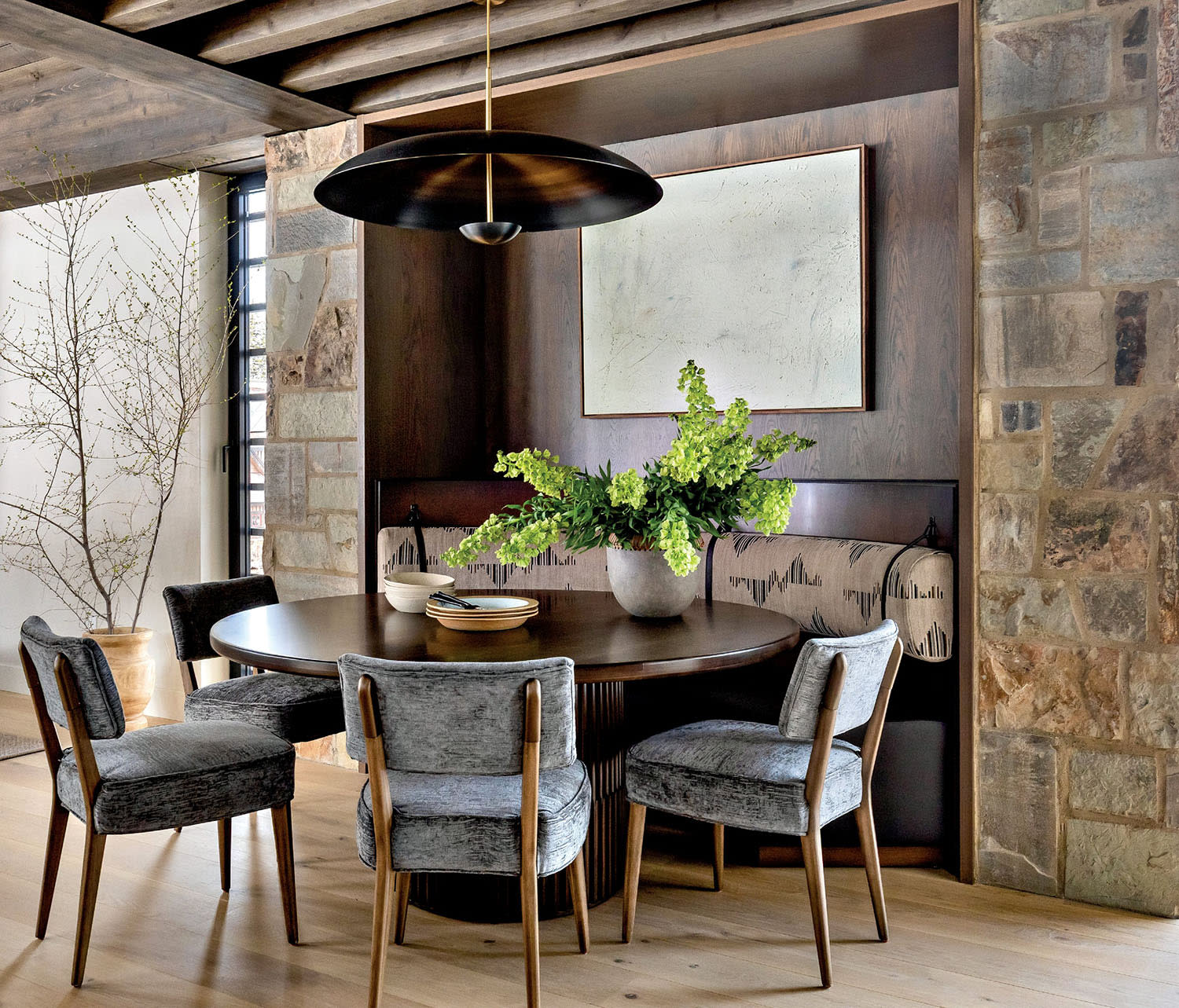 Cozy dining area with a round table, gray upholstered chairs, and a vase of greenery. Rustic stone walls, antler decor, and a modern light fixture.