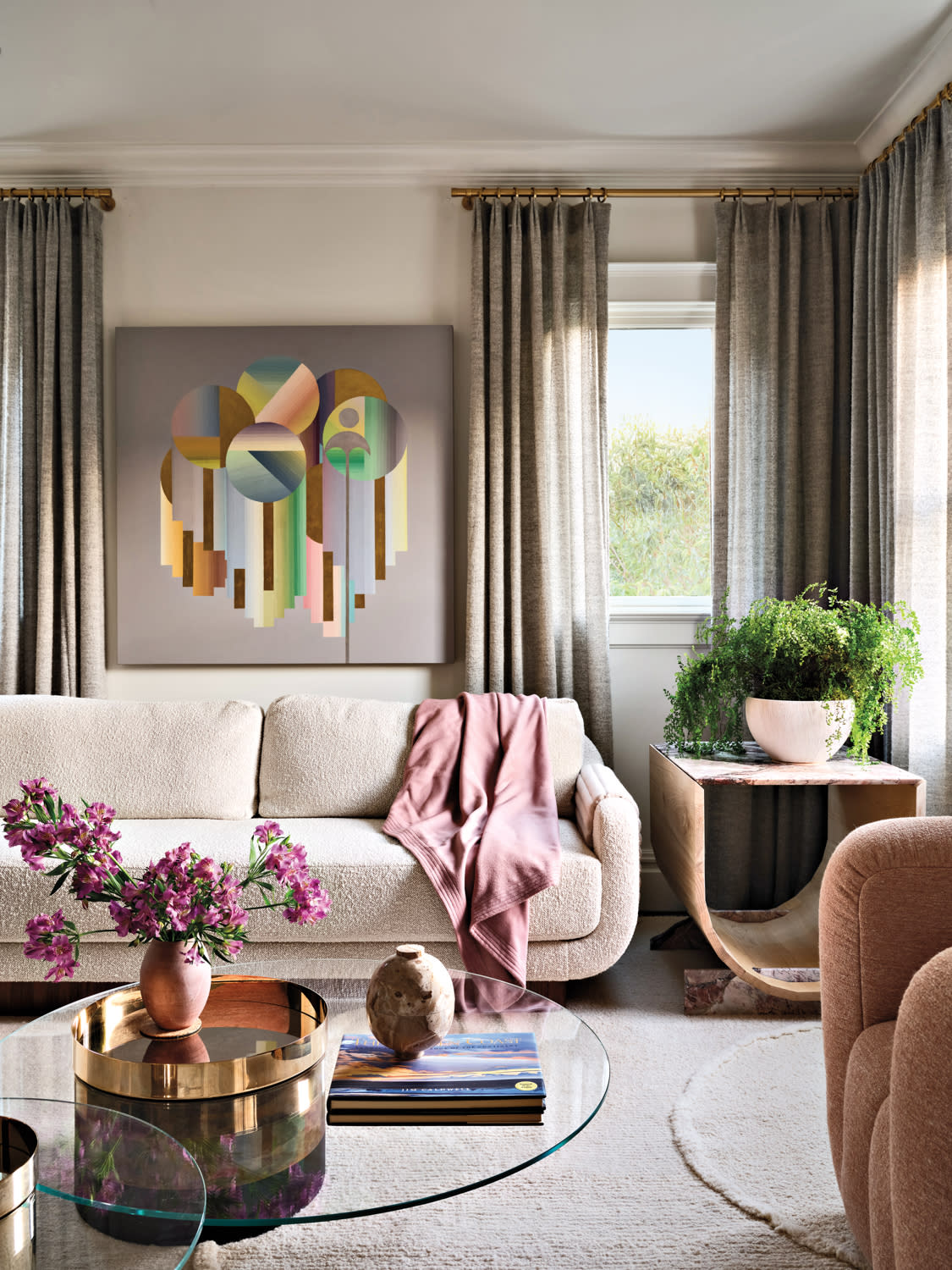 Stylish living room with a cozy cream sofa, colorful abstract art, and a pink throw. Glass coffee table holds flowers and decor; lush potted plant nearby.