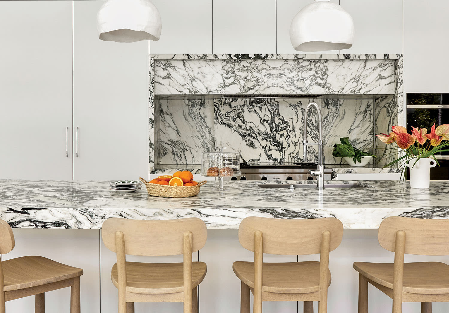 A modern kitchen with a white and black marble countertop and backsplash, wooden stools, and white pendant lights. Fresh flowers and fruit add a vibrant touch.