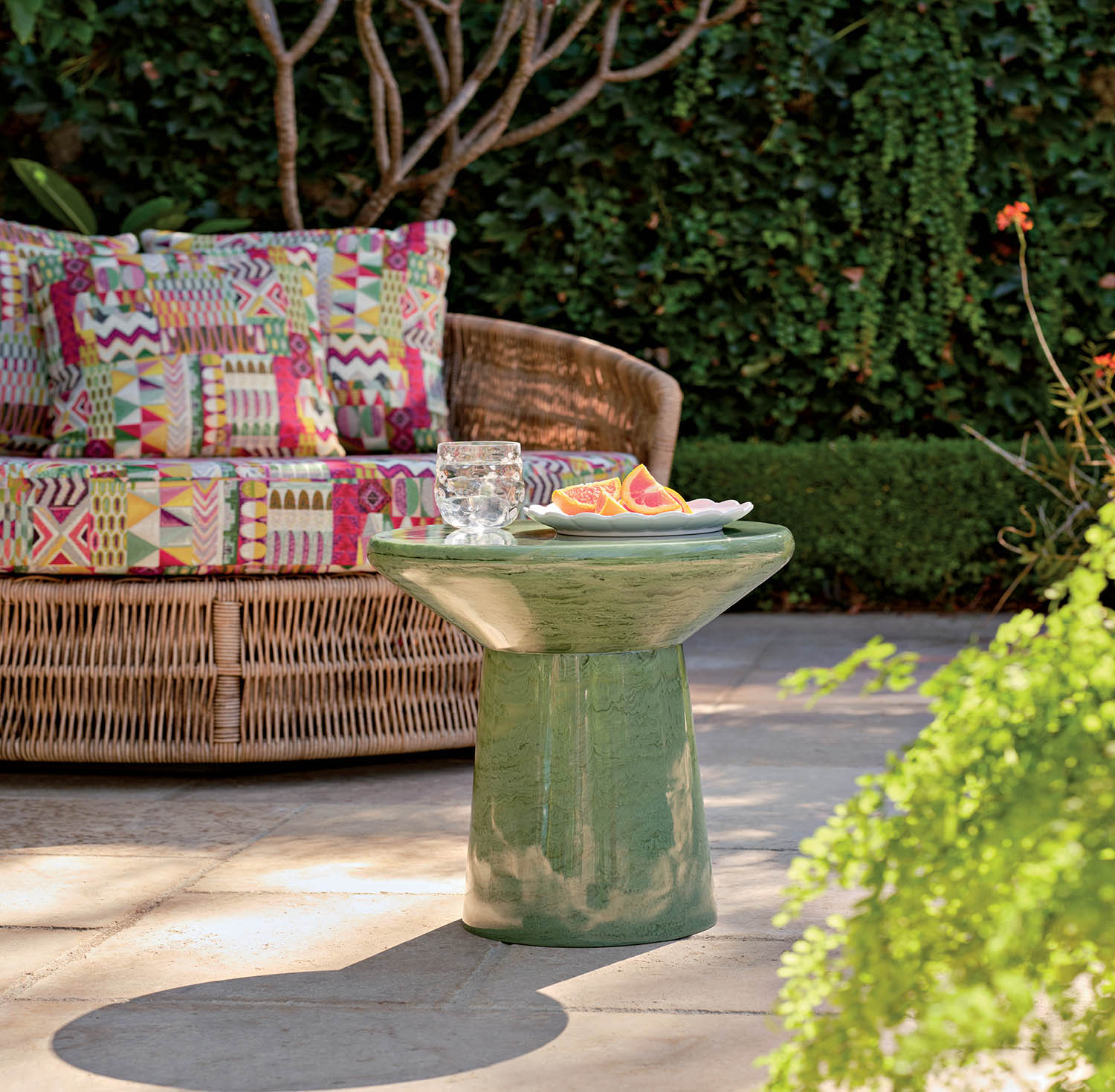 A vibrant patio scene with a wicker loveseat adorned with colorful patchwork cushions. In the foreground, a green ceramic table holds a glass of water and a plate of sliced fruit.