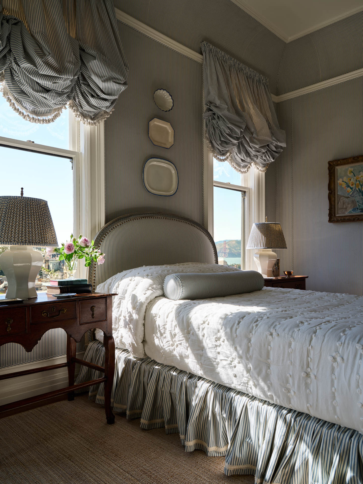 Elegant bedroom with a vintage feel, featuring a striped bedspread and pillows, ornate draped curtains, wooden nightstands, and delicate lamps.