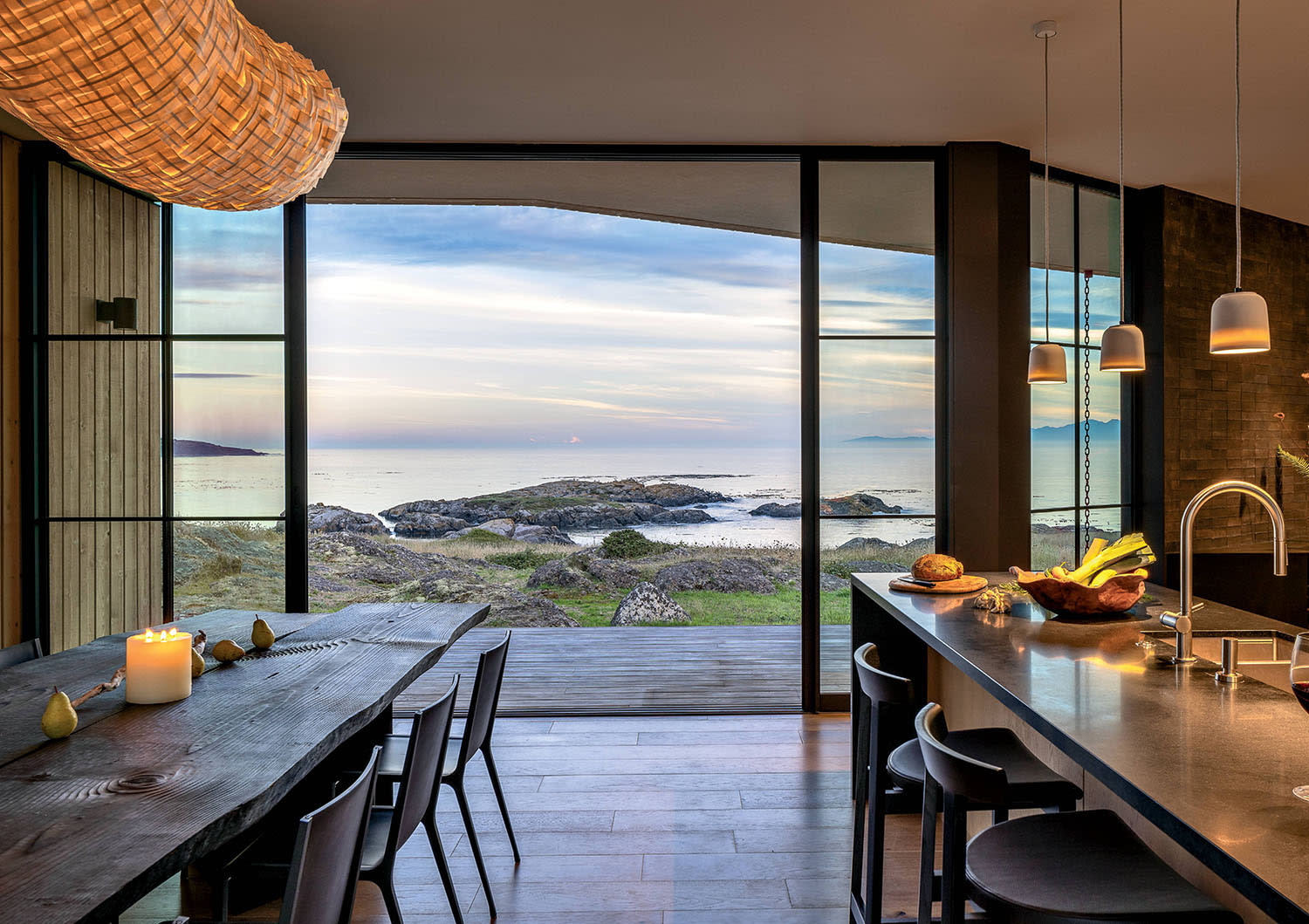 A wood dining and chairs set up parallel to a kitchen island, both near glass doors looking outside.