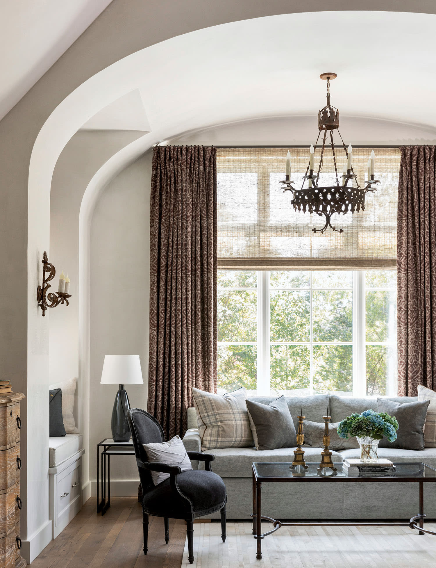 Elegant living room with arched ceiling and large window, featuring a gray sofa, patterned curtains, black armchair, and glass coffee table under a chandelier.