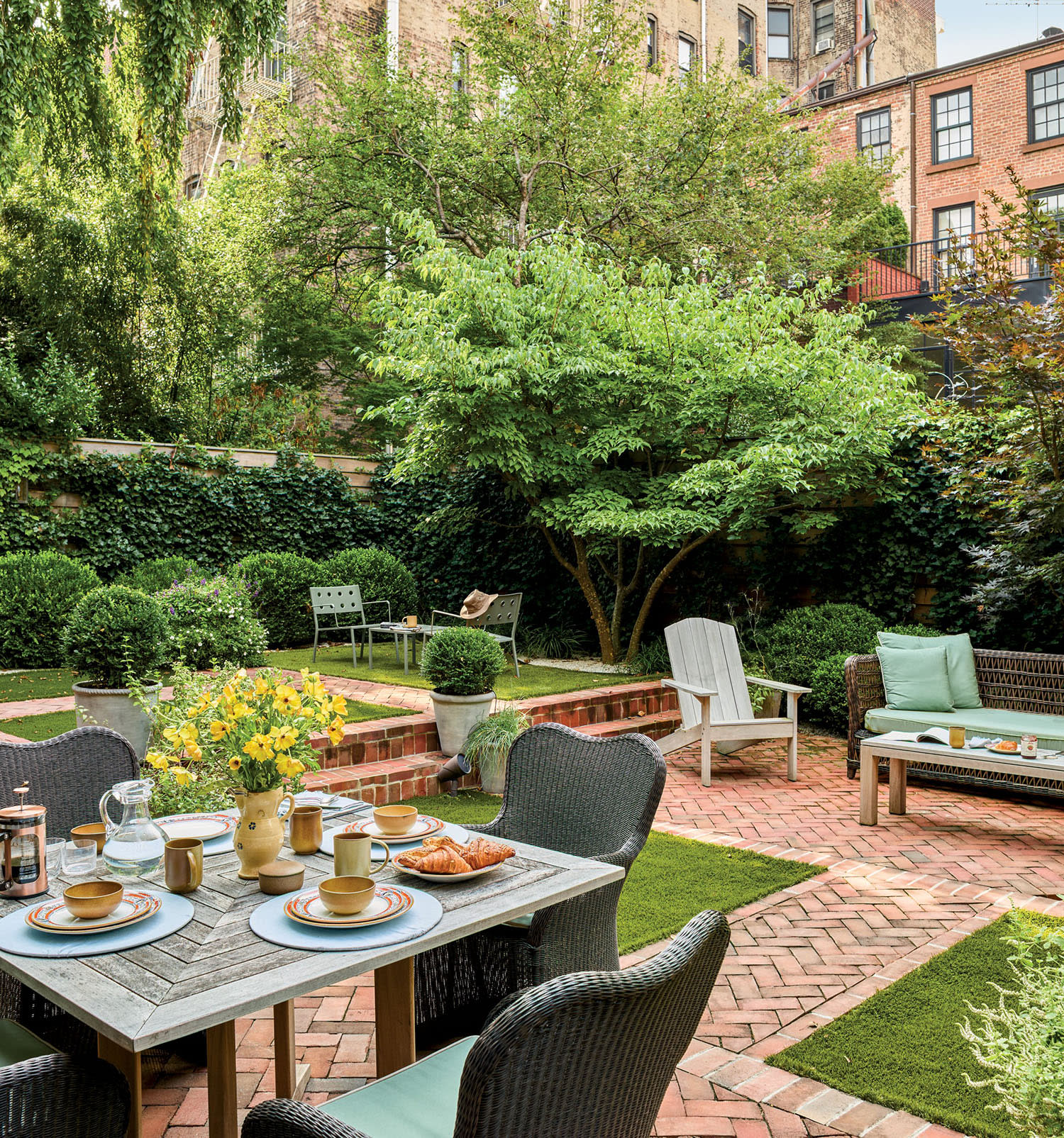 Charming urban garden with brick patio featuring a dining table set for breakfast, surrounded by lush greenery, potted plants, and cozy seating areas.