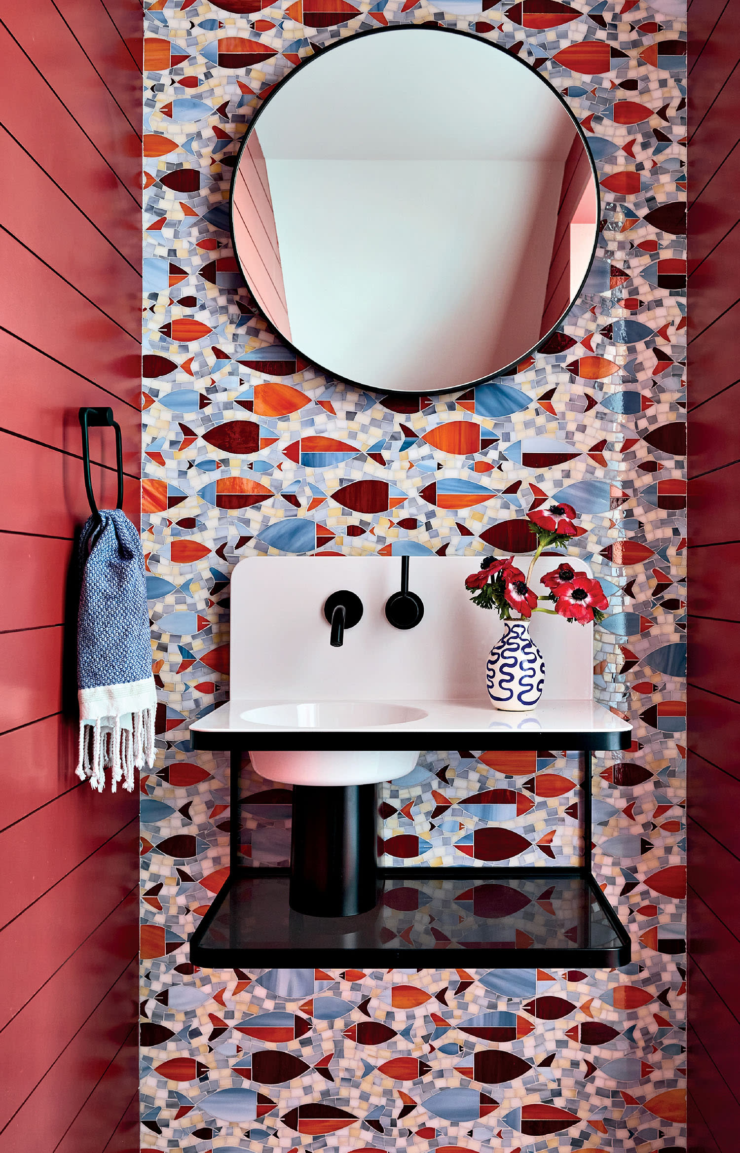 Bathroom with bold patterned tiles featuring geometric shapes in reds and blues. A round mirror and modern sink are accented by red walls and a vase with red flowers.