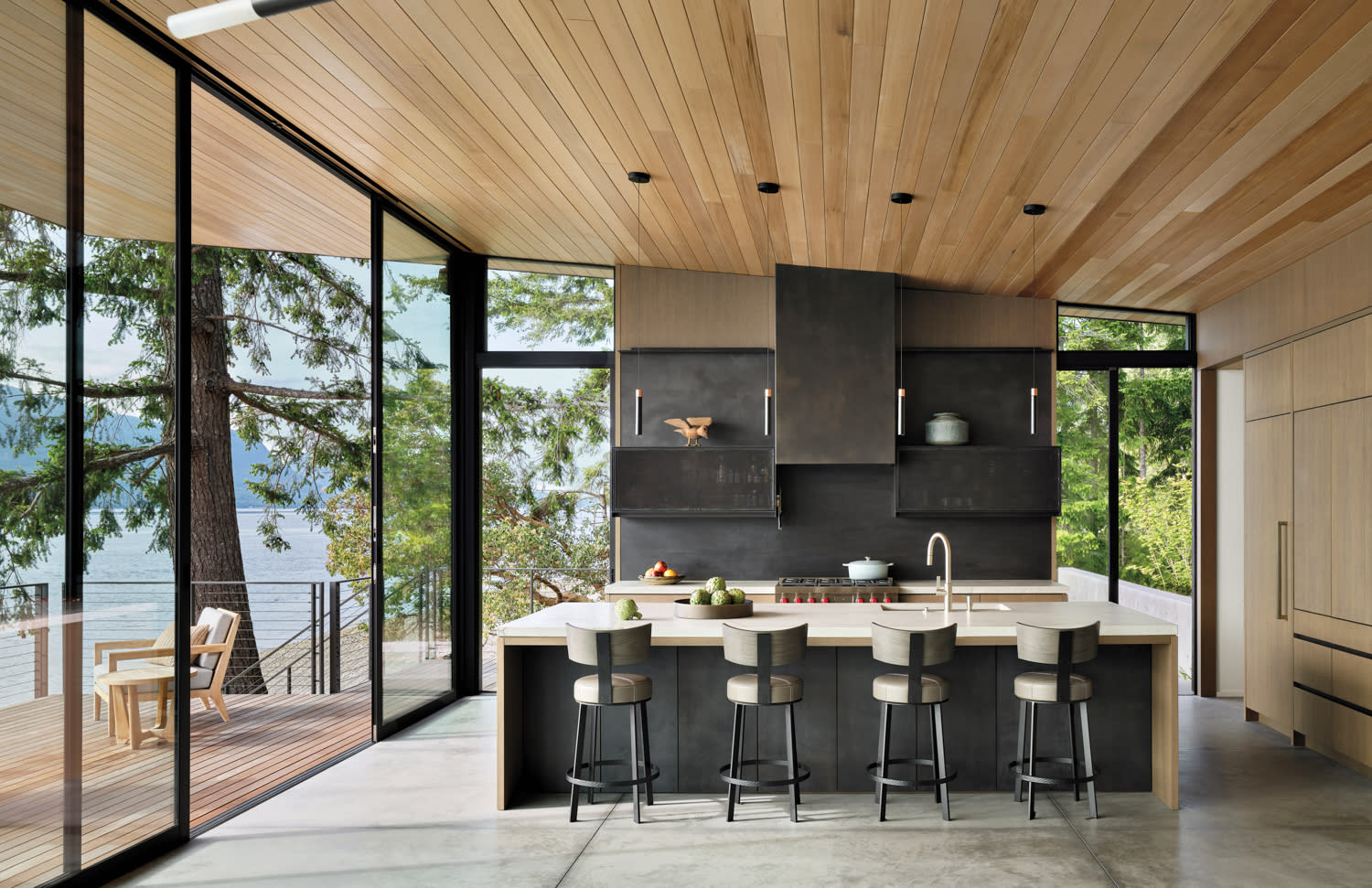 Modern kitchen with wooden ceiling and large glass windows showcasing a scenic lake view. Features a sleek island with four barstools and open shelves.