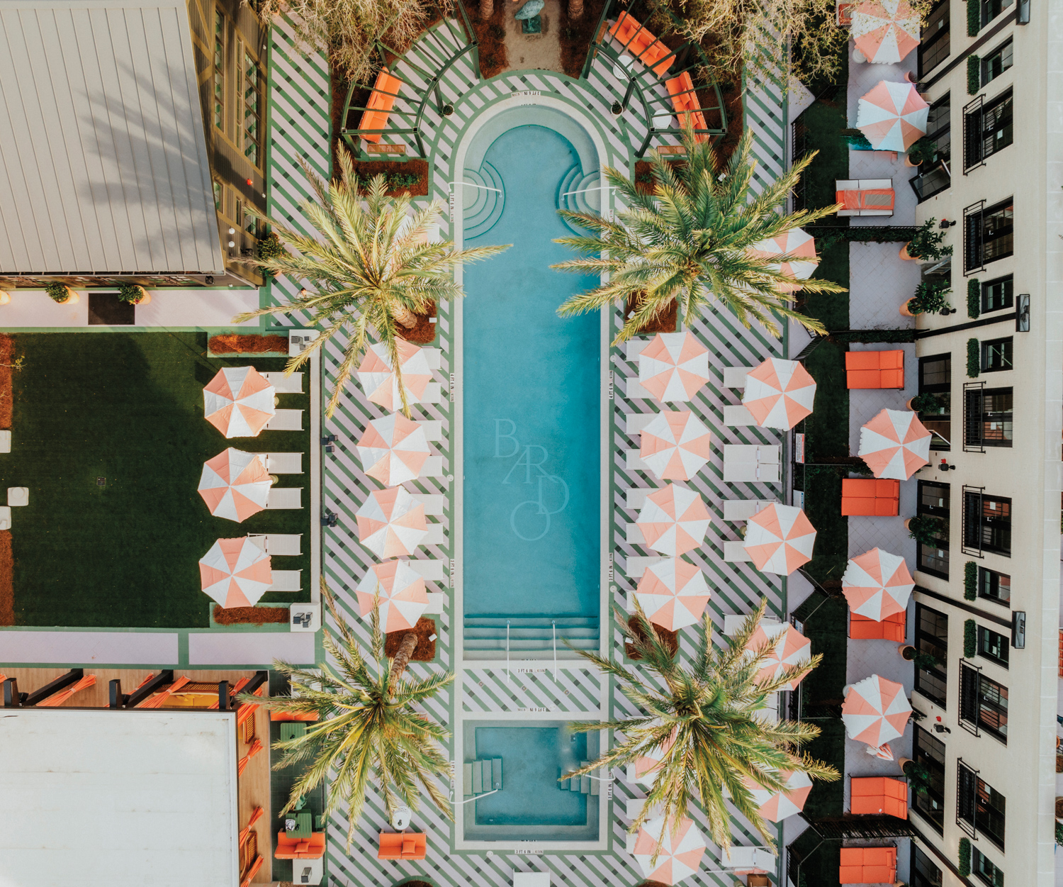 Birds-eye view of long, thin hotel swimming pool lined with pink-and-white umbrellas anod white chaises. 