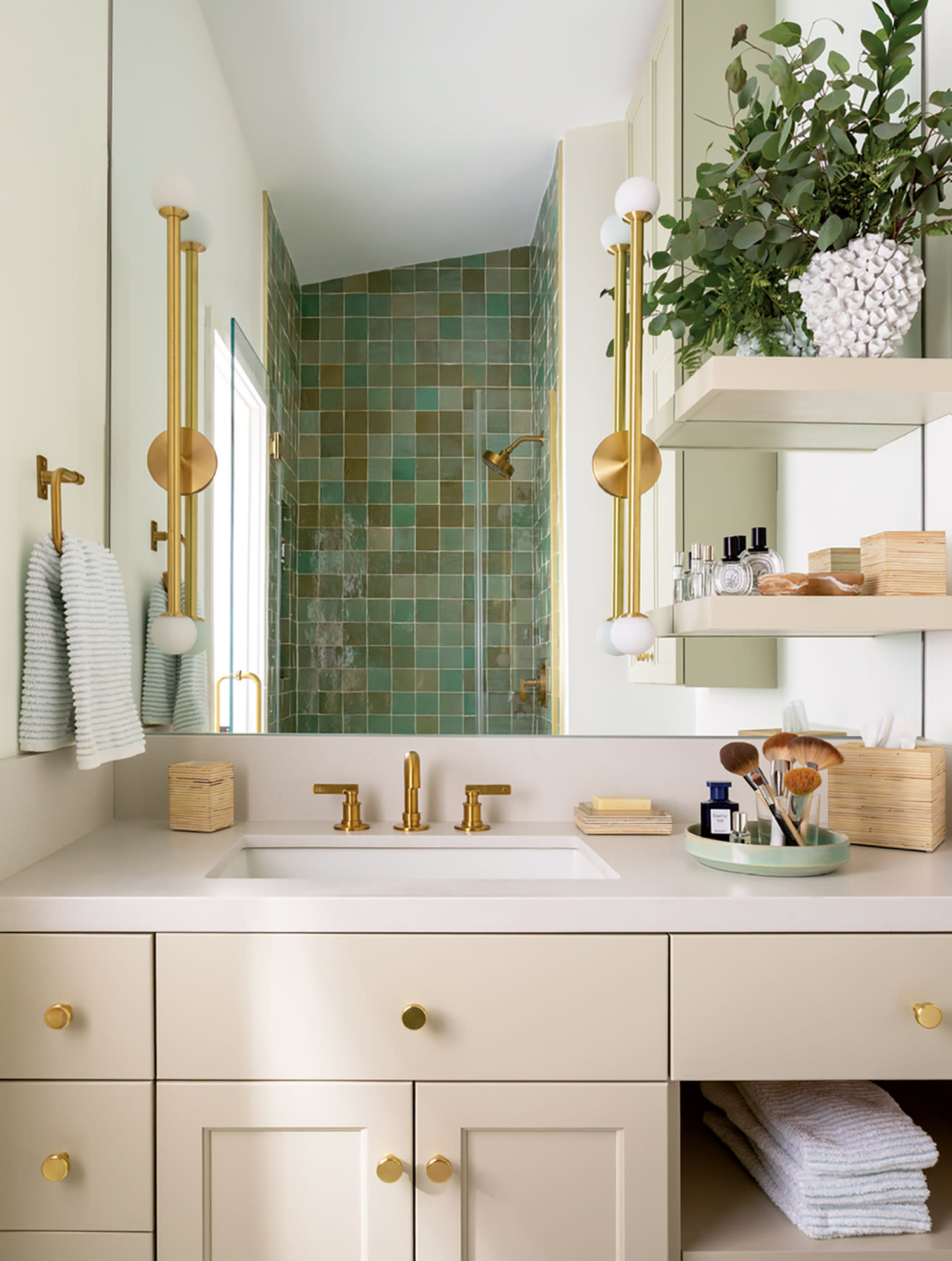 Elegant bathroom with beige cabinets and gold fixtures. Features a green-tiled shower, white countertop, plants, and neatly arranged towels. Bright and organized.