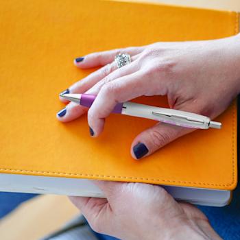 Main de femme tenant un stylo Nash deluxe blanc à grip violet posé sur un carnet orange