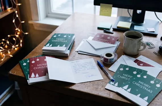 Desk with a stack of Christmas cards, a handwritten card, envelopes, stamps and a ticked-off list of names