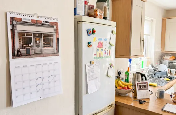 Printed wall calendar next to the fridge in a family kitchen, with a desk calendar on the counter