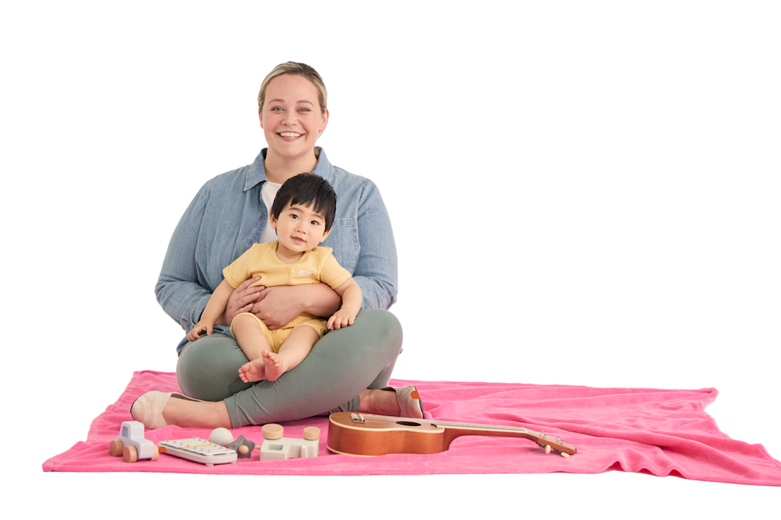 Babysitter holding a baby, both smiling while sitting on a mat with toys