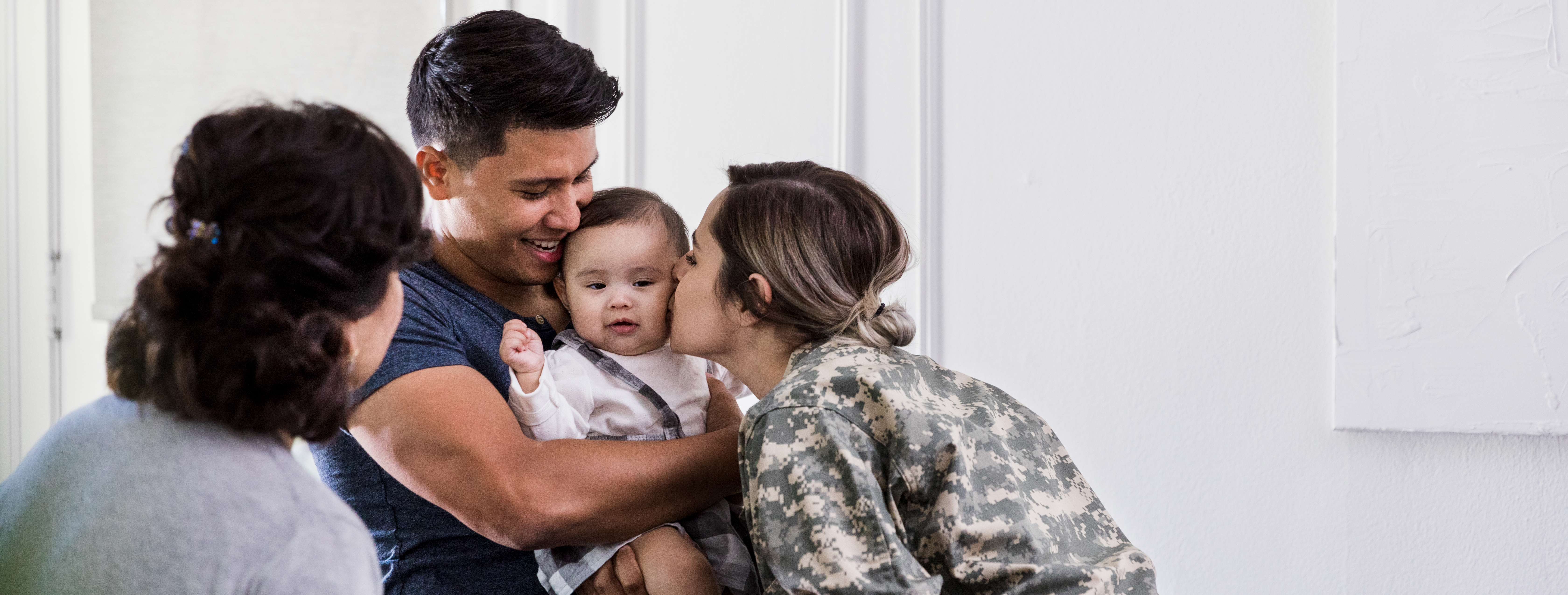 Nanny holding a baby as their military mom kisses their cheek.