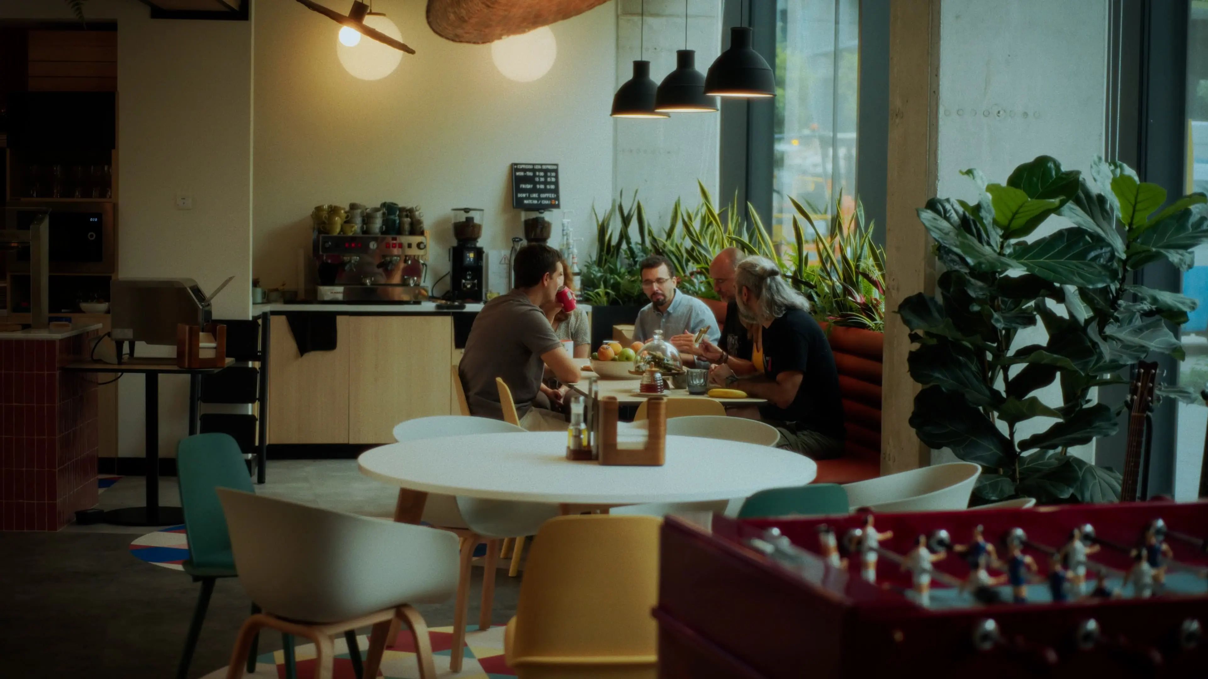 Group of people having a meeting in a modern café with plants, pendant lights, and coffee equipment in the background.