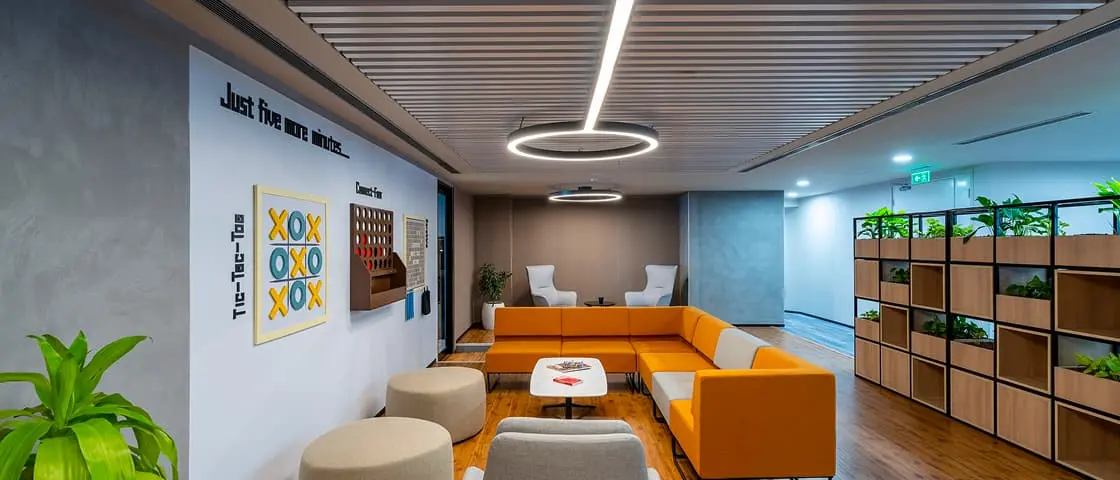 Modern office lounge with orange sectional sofa, tic-tac-toe wall art, wooden floor, and plant-filled shelving units.