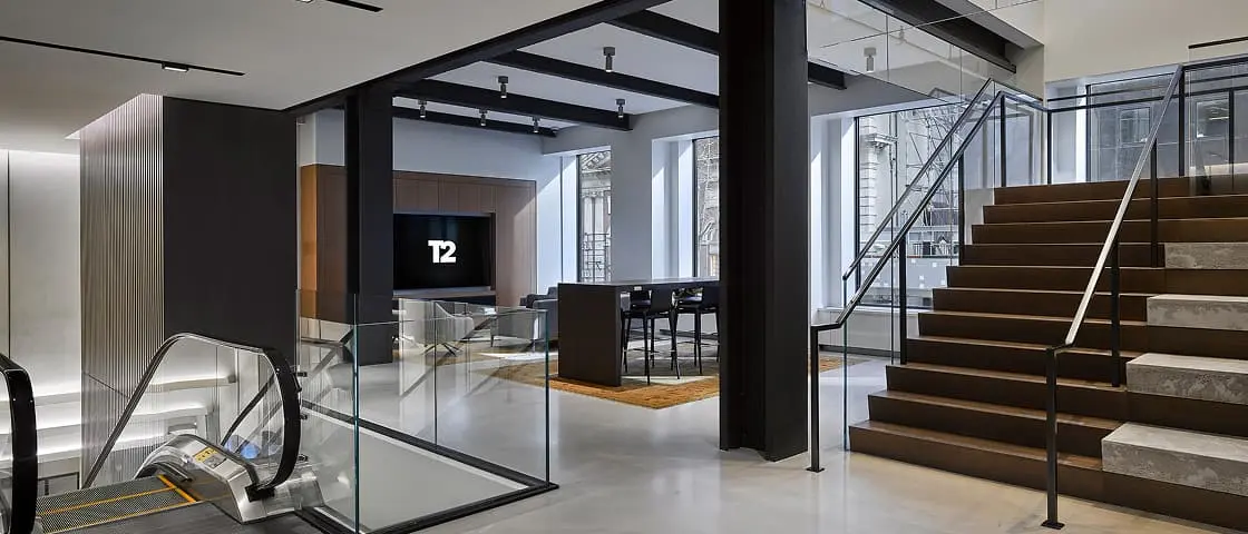Modern office interior with glass staircase, escalator, and "T2" displayed on wall screen in sleek, open space design.