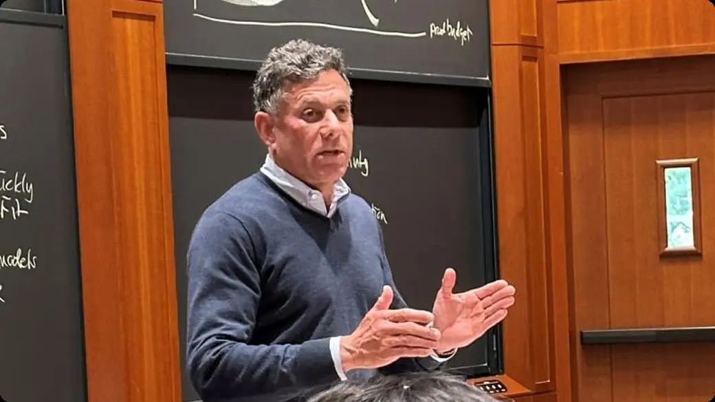 Strauss Zelnick, CEO of Take-Two Interactive Software, Inc. in blue sweater gesturing with hands during presentation in wood-paneled room with blackboard behind.