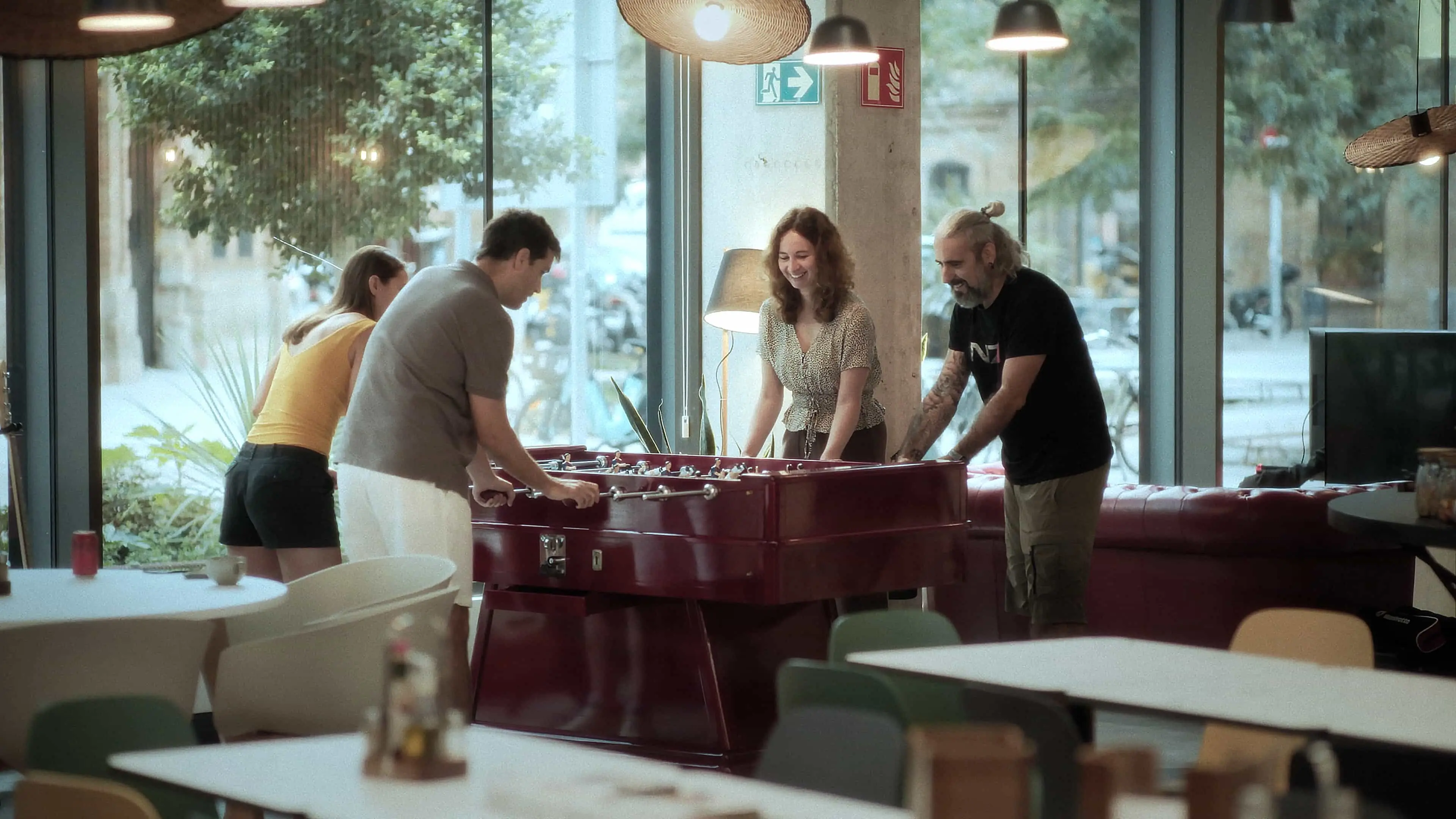 Four people playing foosball at a red table in a modern café with large windows overlooking a street with trees.