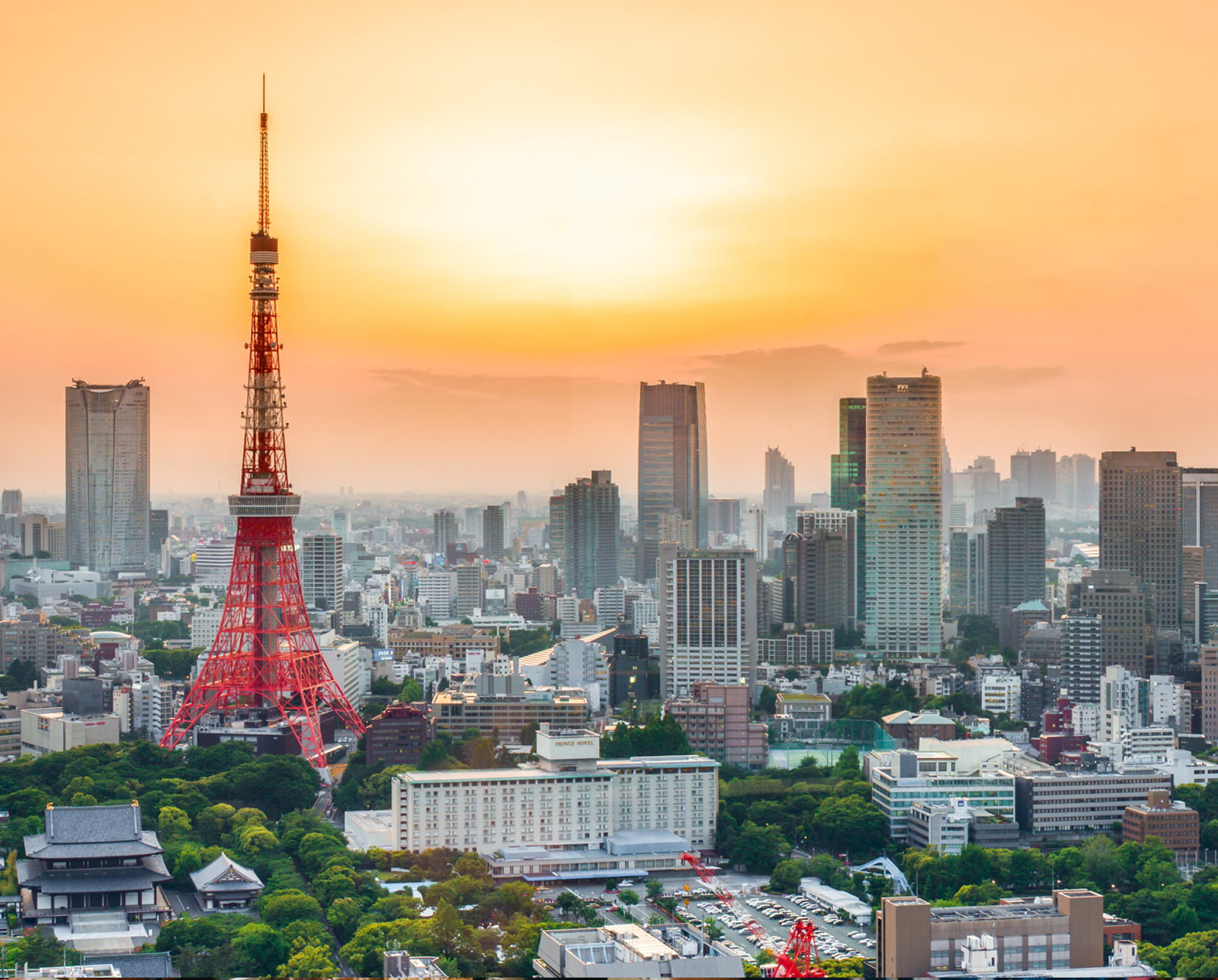 Tokyo Tower stands tall against a sunset, surrounded by city buildings and greenery, showcasing urban Tokyo's skyline.