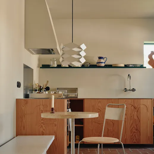 A folded pendant lamp above a round bistro table in Carla Camilla's kitchen, with walls in KABRIC Mushroom and hand-carved wooden fronts.
