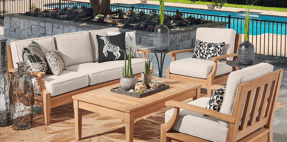 Outdoor patio with beige cushioned wooden furniture, animal print pillows, plants, and a pool in the background.