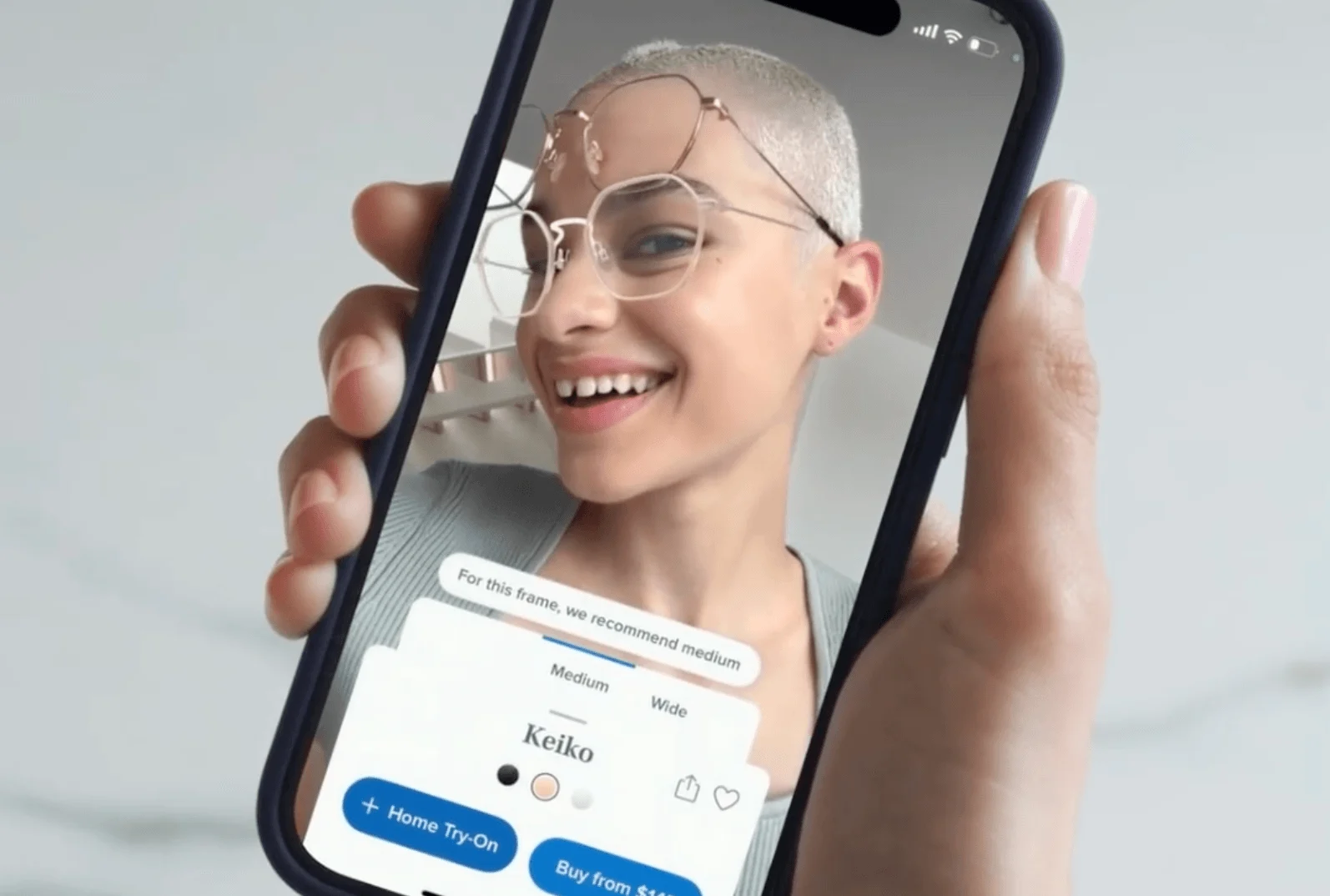Hand holding smartphone showing virtual try-on of eyeglasses with smiling person wearing geometric frames on screen.