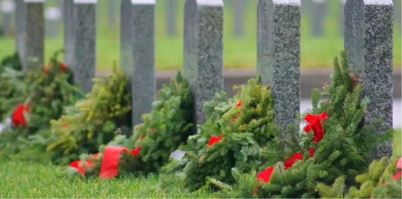 Gray headstones in a row with evergreen wreaths decorated with red bows at their bases on green grass.