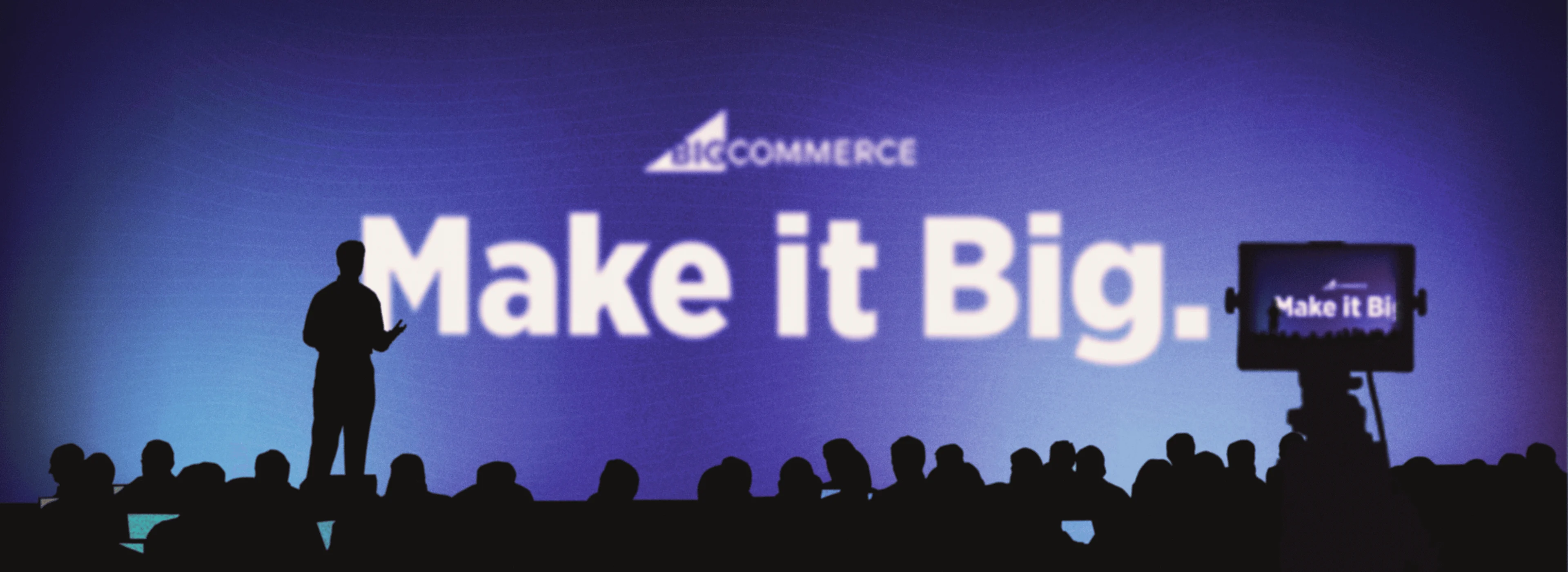 Silhouette of a speaker on stage with "Make it Big" and "BigCommerce" on a blue screen, audience and camera in foreground.