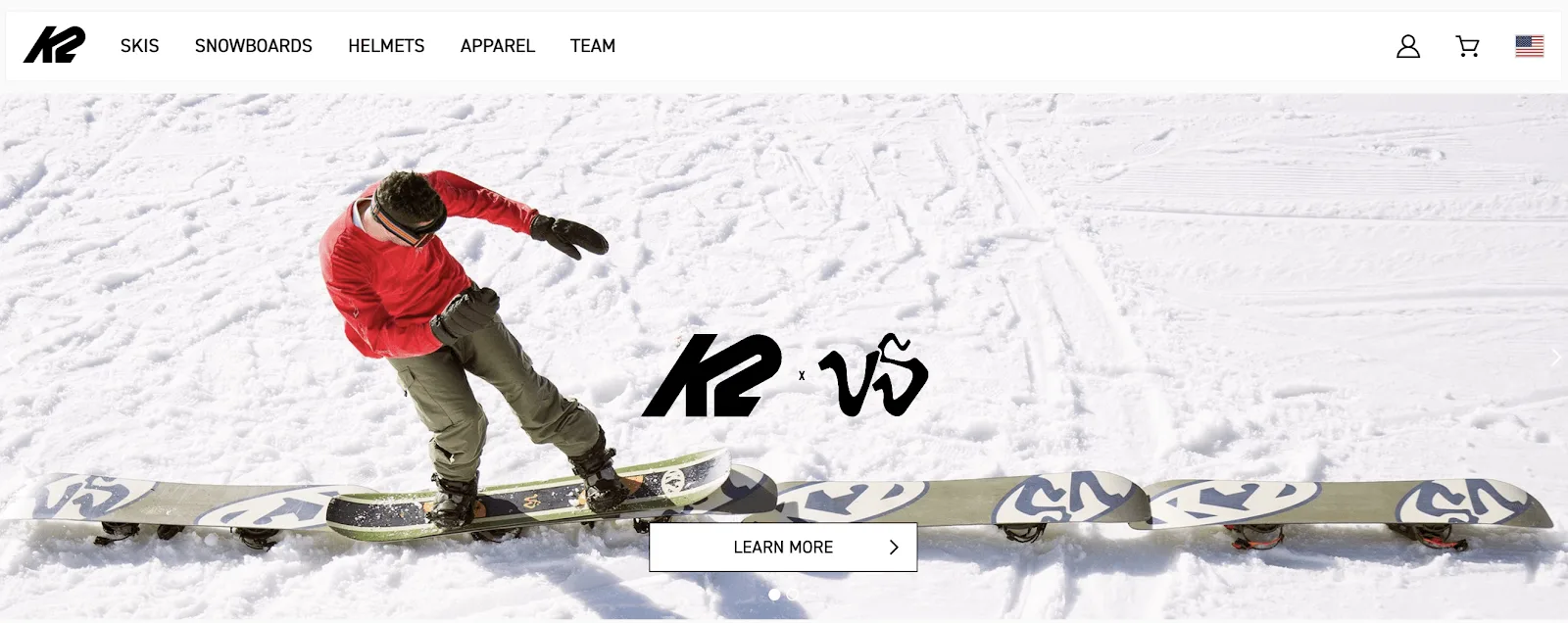 Snowboarder in red jacket and green pants riding on snow, with K2 logo and snowboards in the background.