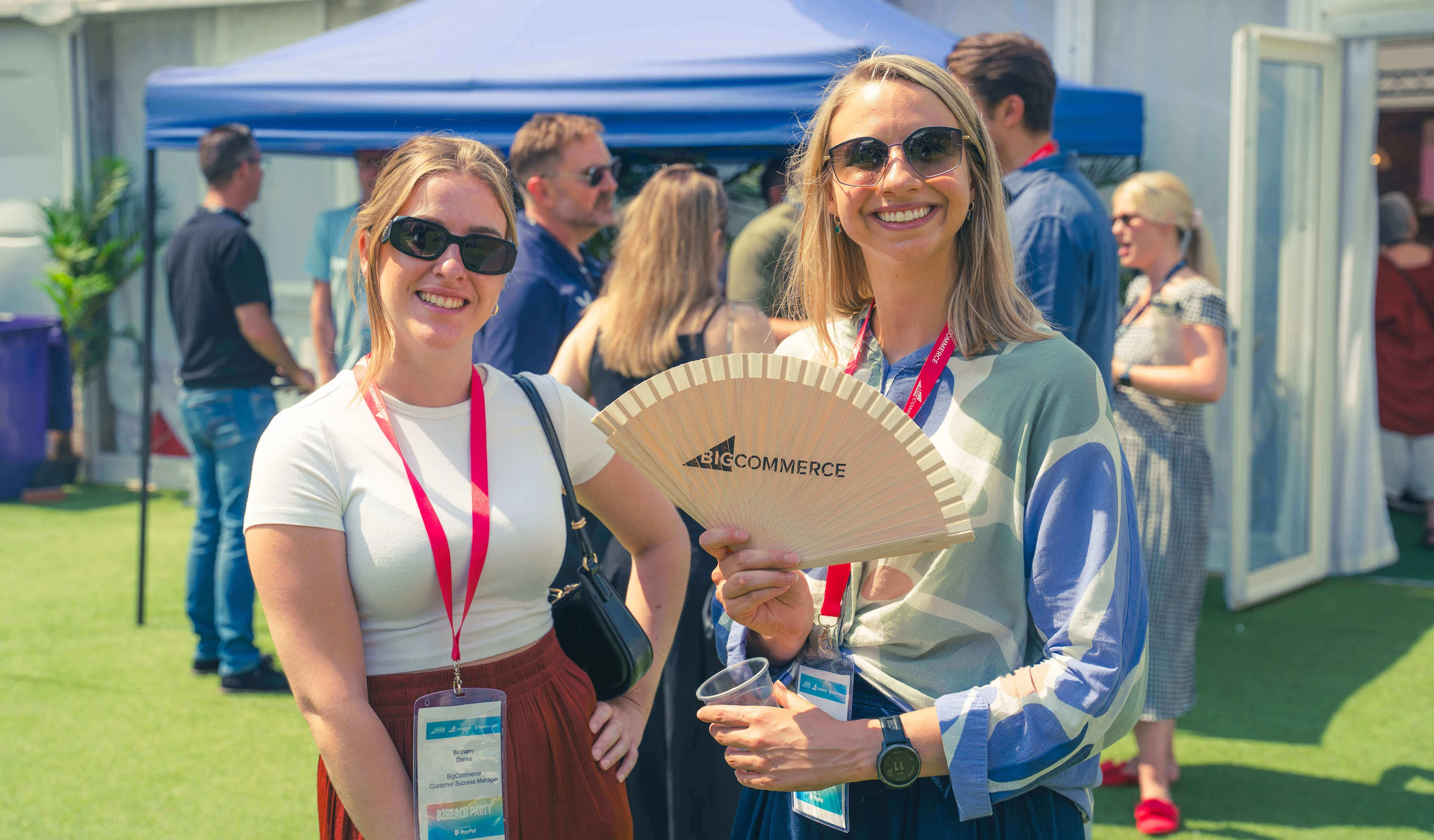 Two smiling women wearing sunglasses at an outdoor event, one holding a fan with the BigCommerce logo.