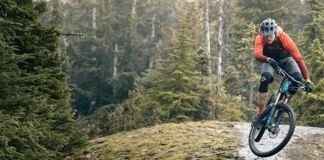 Mountain biker in red and black gear rides a trail through a dense green forest, leaning into a turn on a dirt path.