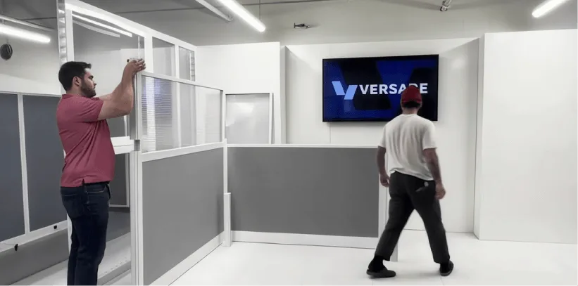 Two people assembling gray and white office cubicle partitions in a modern white office space with Versare logo on screen.
