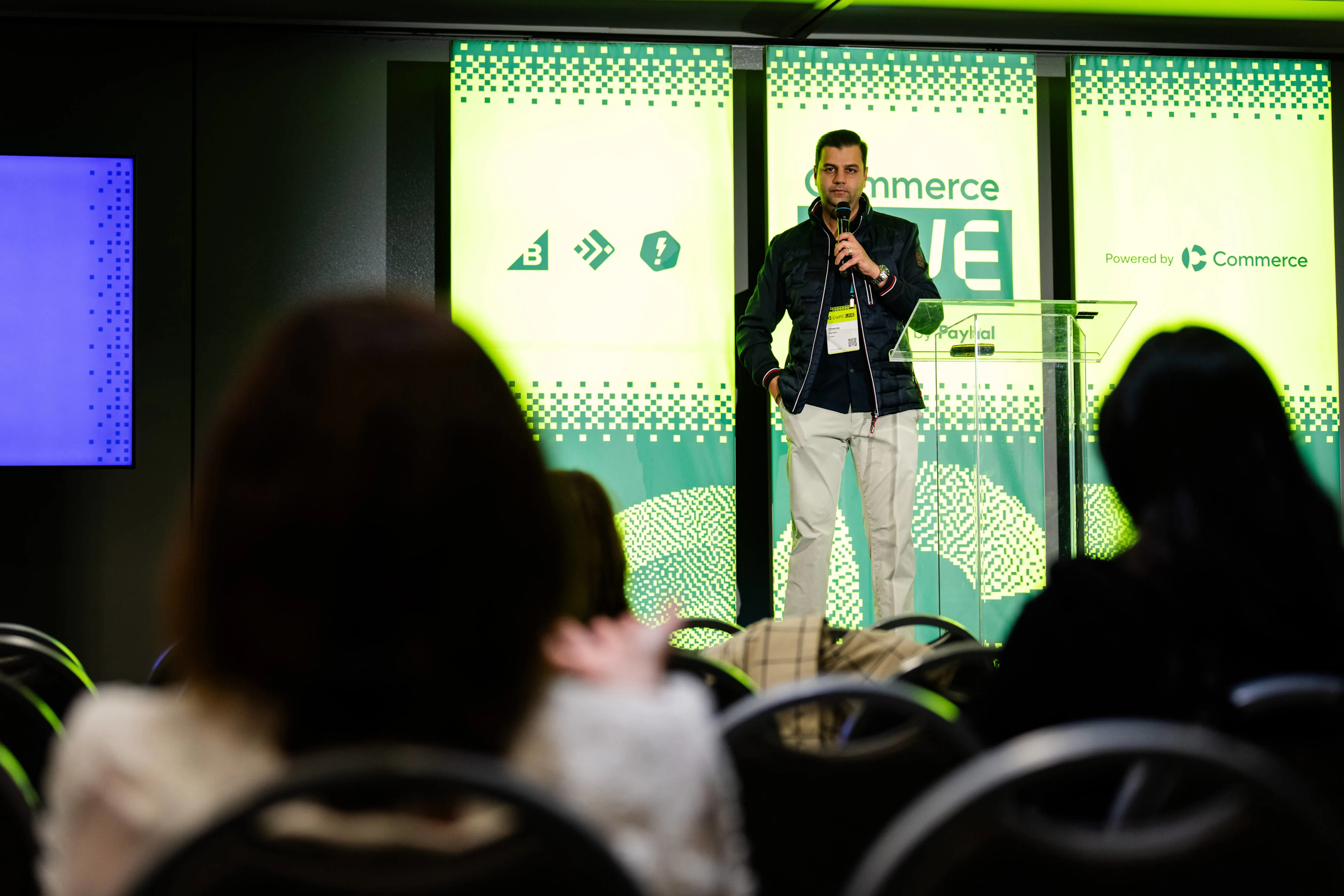 Dheeraj Sareen, Head of Technology Partnerships, Adyen on stage at Commerce Live 2026. Speaker holding a microphone on stage beside a clear podium, green conference backdrop, audience in foreground.