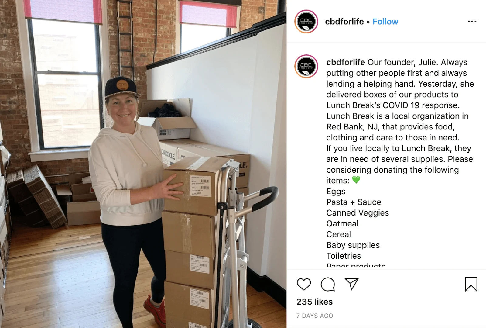 Smiling woman in a white hoodie and hat stands indoors with stacked boxes on a dolly, surrounded by more boxes.
