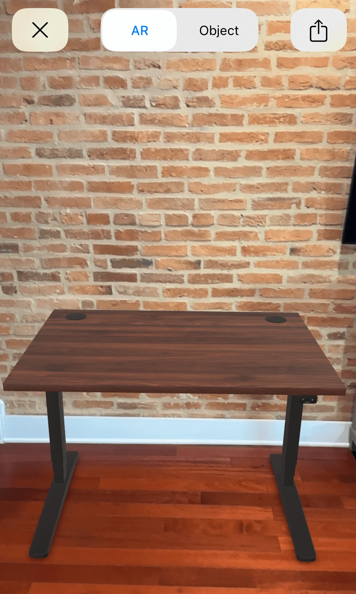A dark wood desk with black legs stands on a reddish wood floor against a brick wall, viewed in augmented reality mode.